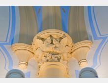 Nahaufnahme eines verzierten Kapitells in einer Kirche oder einem historischen Gebäude, mit floralen Reliefs und gelb-beiger Farbe auf hellem Hintergrund. Der Hintergrund zeigt blau-weiße, bogenartige Malereien an der Decke.