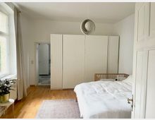 Helles Schlafzimmer mit Holzboden, einem großen weißen Kleiderschrank und einem Bett mit weißer Bettwäsche; daneben steht ein Holzgitterbett. Links ein Fenster mit beigefarbenen Vorhängen und ein kleiner Tisch mit einer Pflanze. Im Hintergrund ist eine geöffnete Tür zu einem gefliesten Raum zu sehen.
