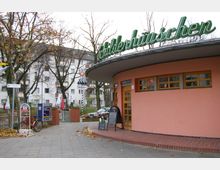 Ein kleines, rundes Gebäude mit rosa Fassade und grüner Leuchtschrift „Schilderhäuschen“ auf dem Dach; im Vordergrund ein Gehweg mit Fahrradständer und Werbetafel, im Hintergrund Bäume und Wohnhäuser.