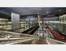 Moderne Bahnhofshalle mit mehreren Ebenen, durchzogen von Rolltreppen und Säulen. Links und rechts befinden sich verglaste Wände, unten verlaufen Bahngleise mit einem roten Regionalzug, dazu sind Anzeigetafeln und Bahnsteigschilder sichtbar.