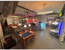 Industriell gestalteter Innenraum mit weiß gestrichenen Ziegelwänden, offener Decke und sichtbaren Metallträgern. Der Raum ist mit einer Mischung aus Holztischen, Stühlen und modernen Leuchten ausgestattet, ergänzt durch farbige Neonlichter und Metallgeländer.