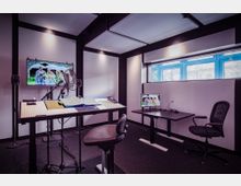 Tonstudio mit schallgedämpften Wänden, einem hohen Tisch mit Mikrofon, Drehstuhl und Monitor mit Filmszene. Im Hintergrund ein Fenster mit blauen Rahmen und ein zweiter Monitor auf einem Schreibtisch.
