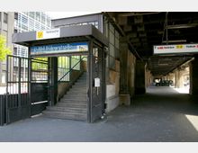 Eingang zum U-Bahnhof Bülowstraße in Berlin, mit einer Treppe, die zur Station der Linie U2 führt. Das Gebäude befindet sich unter einer Brücke, deren Stahlkonstruktion sichtbar ist, und im Hintergrund sind moderne Bürogebäude und parkende Autos zu sehen.