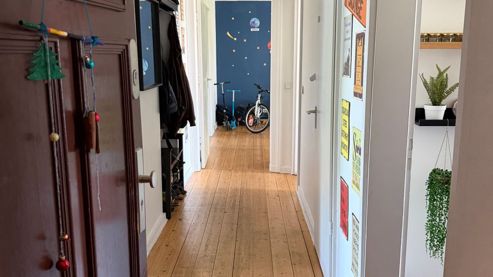 Langer, schmaler Wohnungsflur mit Holzdielenboden, weißen Wänden und einer einzelnen Deckenlampe. Links sind Garderobe und Spiegel neben einer dunklen Eingangstür, rechts ein Türrahmen mit Wandpostern und Regal mit Zimmerpflanzen; am Ende öffnet sich der Flur zu einem Raum mit dunkelblauer Wand und Fahrrad.
