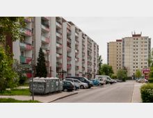 Wohnsiedlung mit mehreren mehrstöckigen Gebäuden; links sind Wohnblöcke mit Balkonen in Weiß und Rot zu sehen, rechts ein höheres Gebäude in Beige. Im Vordergrund befinden sich Müllcontainer, parkende Autos und ein Straßenabschnitt mit Beschilderung ("Ringenwalder Straße"). Im Hintergrund sind Bäume und ein Supermarktgebäude mit den Logos von REWE und PENNY erkennbar.