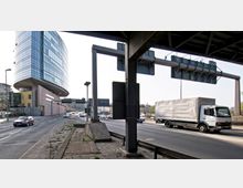 Eine städtische Straße mit mehreren Fahrspuren und Fahrzeugen, darunter PKWs und ein LKW. Im Hintergrund ein modernes Bürogebäude mit gebogener Glasfassade, daneben Verkehrsschilder und eine Brücke im oberen Bildbereich.