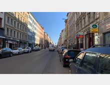 Straßenszene in einer urbanen Umgebung mit mehrstöckigen Altbauten auf beiden Seiten, teils in hellen Farben gestrichen. Autos parken entlang der Straße, und Geschäfte mit Werbeschildern befinden sich im Erdgeschoss der Häuser.