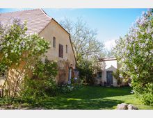 Ein ländlicher Hof mit einem beige verputzten Gebäude mit roten Ziegeldach, kleinen Fenstern und Holztüren. Der Hof ist von Fliederbüschen, anderen Pflanzen und einer Rasenfläche umgeben, mit freiem Himmel im Hintergrund.