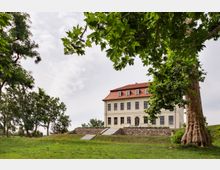 Herrenhaus mit heller Fassade und rotem Walmdach, umgeben von einer Rasenfläche und Bäumen. Das Gebäude steht auf einem erhöhten Gelände mit einer Steintreppe und einer Mauer.