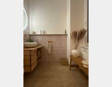 Helles Badezimmer mit halbhoher, beige gefliester Wand und grauem Steinboden. Links steht ein Holzwaschtisch mit runder Aufsatzschale und goldfarbenem Wasserhahn, rechts eine Holzbank neben einem Fenster mit weißer Vase und Trockenpflanzen.