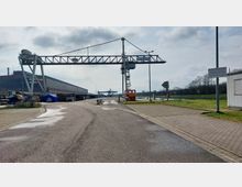 Industriegelände mit Hafenbereich, breiter asphaltierter Weg neben einem Wasserkanal. Im Vordergrund ein großer Hafenkran, links ein großes Lagergebäude und Baugeräte, rechts ein kleines Containergebäude sowie eine Wiese und Schilder mit Warnhinweisen.