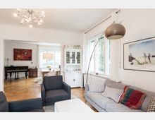 Wohnzimmer mit heller Einrichtung, bestehend aus einer grauen Couch, einem weißen Couchtisch und modernen Lampen. Im Hintergrund ein Essbereich mit großem Fenster, einem Kronleuchter und einem Klavier; Parkettboden zieht sich durch beide Räume.