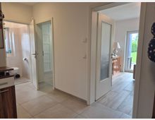 Heller Flur in einer Wohnung mit grauen Bodenfliesen und weißen Wänden; links führt eine offene Tür in ein weiß gefliestes Badezimmer. Rechts öffnet sich der Flur zu einem Wohnraum mit Glastür, Holzmöbel und einer großen Terrassentür mit Blick ins Grüne.