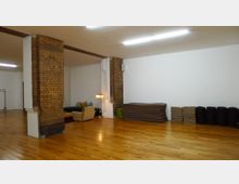 Ein heller Raum mit Holzboden und freistehenden Backsteinpfeilern. An einer weißen Wand sind gestapelte Matten und Kissen zu sehen, während auf der linken Seite Sitzmöbel mit einer Stehlampe stehen.