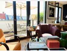 Wohnzimmer mit Zugang zu einer Terrasse, geprägt von bodentiefen Fenstern, die viel Licht hereinlassen. Innen befinden sich farbige Polstermöbel, ein Couchtisch mit Büchern und Dekorationen, während draußen auf der Terrasse ein Sonnenschirm und Sitzmöbel stehen.