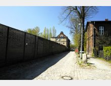 Kopfsteinpflasterstraße mit einer hohen Ziegelmauer auf der linken und Backsteinhäusern auf der rechten Seite. Im Hintergrund ist ein mehrstöckiges Gebäude mit Giebel und umliegenden Bäumen sichtbar, bei klarem Himmel und Tageslicht.