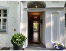 Eingangsbereich eines hellen Hauses mit weißer Fassade und Strukturputz. Geöffnete Tür mit Blick durch den Flur auf eine Terrasse; Blumenkübel mit Margeriten und violetten Blumen flankieren den Eingang. Ein Kronleuchter ist im Inneren sichtbar.