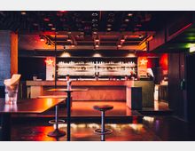 Eine moderne Bar mit einem langen Tresen aus Holz, dahinter Regale mit Gläsern und Flaschen, beleuchtet von warmem Licht. Im Vordergrund stehen runde Tische mit Hockern, die Szenerie ist in rötlichem Licht gehalten, mit dekorativen Deckenleuchten und einem roten Leuchtstern an der Wand.