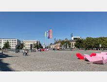 Weitläufiger öffentlicher Platz mit Pflastersteinen, umgeben von historischen Gebäuden und Bäumen. Im Vordergrund befinden sich moderne rosa- und rotfarbene Sitzbänke sowie Fahrradständer, während bunte Fahnen und ein Zugang zur U-Bahn zu sehen sind.