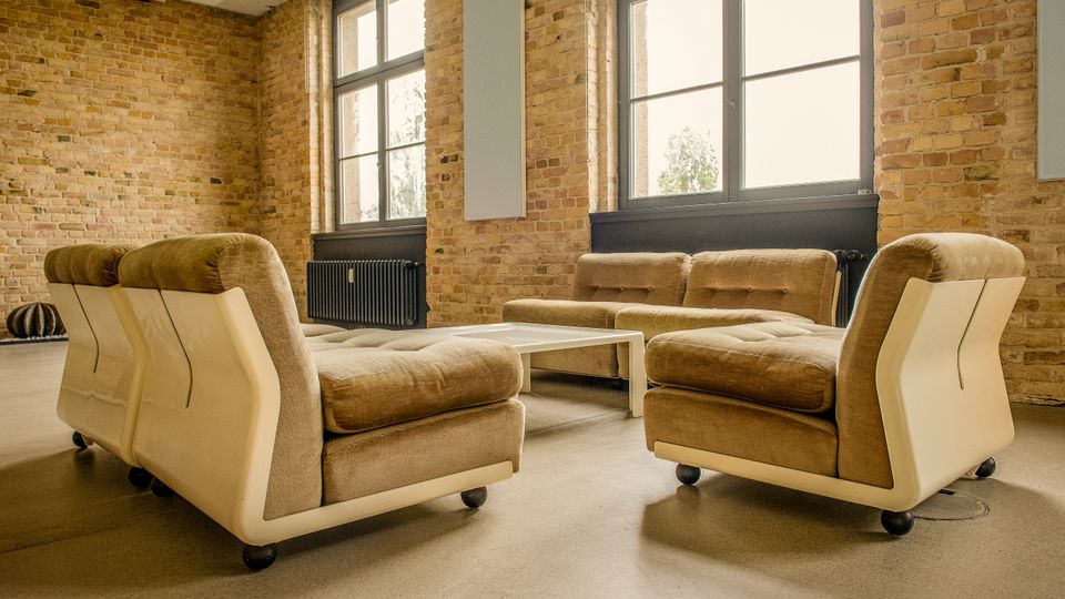 Loftartiger Innenraum mit unverputzten Ziegelwänden, großen Sprossenfenstern und Heizkörpern unter den Fenstern. Im Vordergrund stehen beige Polstersofas auf Rollen um einen niedrigen weißen Couchtisch auf glattem Betonboden.