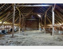 Innenraum eines alten Dachbodens mit Holzbalkenkonstruktion und bedachtem Giebel. Der Boden ist mit Heu bedeckt, und durch kleine Fensteröffnungen fällt Tageslicht in den Raum.