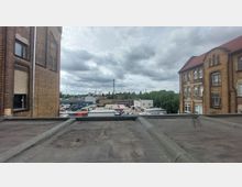 Blick von einem Flachdach auf einen Gewerbehof mit Containern, Lagerhallen und geparkten Fahrzeugen. Im Vordergrund sind zwei Backsteingebäude mit großen Fenstern, im Hintergrund eine Mischung aus Bäumen, Industriebauten und einem hohen Sendemast unter bewölktem Himmel.