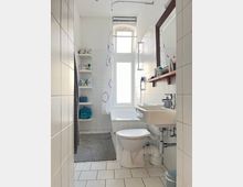 Helles Badezimmer mit weiß gefliesten Wänden und Boden; ausgestattet mit einer Badewanne mit Duschvorhang, einem Waschbecken, einem WC und einem großen Fenster, das Tageslicht hereinlässt. An der Wand sind offene Regale mit Handtüchern und Pflegeprodukten.