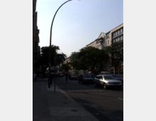 Straßenansicht in einer städtischen Umgebung mit mehrspuriger Fahrbahn, auf der Autos fahren. Rechts säumen Gebäude mit mehreren Stockwerken und Geschäften die Straße, während links ein Gehweg und Bäume sichtbar sind.