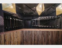 Ein moderner Garderobenbereich mit dunklen Wänden, einer Theke aus vertikal gestreiftem Holz und verstellbaren Haken an Metallrahmen. Die Decke ist perforiert, mit geometrischen Hängelampen aus Metallgewebe.