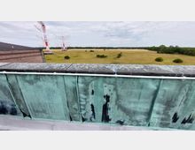 Blick von einem kupfergedeckten Dach mit sichtbarer Gravur „Heinrich Kurz, Berlin“ auf eine große, offene Wiesenlandschaft. Im Hintergrund sind rot-weiße Antennenmasten zu sehen, umgeben von Bäumen und einem bewölkten Himmel.