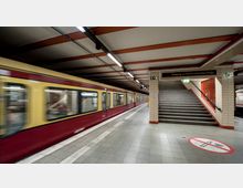 Ein U-Bahnhof mit einer gelb-roten S-Bahn, die auf einem Gleis einfährt. Rechts befinden sich eine Treppe mit einem Schild „Ausgang zur Garten- u. Bernauerstraße“ sowie Wandfliesen in Beige; auf dem Boden ist ein großes Rauch- und Essverbotssymbol sichtbar.