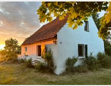 Ein kleines, weiß gestrichenes Landhaus mit einem roten Ziegeldach, umgeben von Sträuchern und Bäumen, bei Sonnenuntergang. Der Vorderbereich zeigt geöffnete Fensterläden, einen Holzstuhl und eine grüne Wiese im Vordergrund.