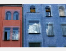 Fassade eines modernen Wohngebäudes mit zwei unterschiedlich farbigen Bereichen: links in Rot und rechts in Blau, beide mit glatten Putzflächen und großen Fenstern, die mit Glasplatten bedeckt sind; die Spiegelung von Wolken ist in den Fenstern sichtbar.
