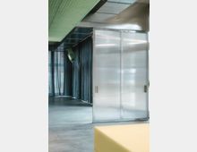 Innenraum mit modernen Designelementen: Eine große Schiebetür aus transparentem geriffeltem Kunststoff mit metallischem Rahmen, schwarzer Vorhang im Hintergrund, polierter Betonboden und eine grüne, perforierte Decke. Im Vordergrund ist eine gelbe Sitzfläche sichtbar.