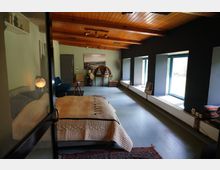 Gemütliches Schlafzimmer mit Holzbalkendecke, einem Bett mit heller Steppdecke, modernem blauen Sofa, und einem antiken Schreibtisch an der Wand. Große Fenster auf der rechten Seite lassen viel Tageslicht herein, während der Boden in einem hellen Blau gehalten ist.