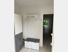 Heller Eingangsbereich mit einer Garderobe bestehend aus einer weißen Wandplatte, Kleiderhaken, Kleiderbügeln und einer kleinen Ablage. Darunter befindet sich eine weiße Sitzbank mit grauer Polsterung und zwei Türen, daneben ein Schrank mit grauer und weißer Oberfläche. Im Hintergrund führt eine Tür zu einem Flur mit Blick auf einen Garten.