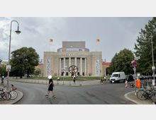 Historisches Gebäude der Volksbühne in Berlin mit klassizistischer Fassade und Säulen, davor ein großer Platz mit einer Skulptur in Form eines stilisierten Zahnrads. Im Vordergrund sind Fußgänger, Fahrräder und parkende Autos auf einer Straße sichtbar.