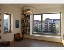 Wohnraum mit Bücherregal, großem Fenster und Stadtblick. Der Raum hat Holzboden, minimalistische Möbel, eine auffällige Hängelampe und zwei kleine Holzkisten auf dem Boden.