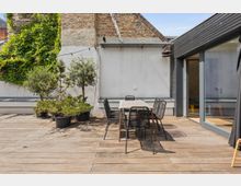 Dachterrasse mit Holzboden, ausgestattet mit einem Tisch und sechs Stühlen aus Metallgeflecht. Im Hintergrund stehen Pflanzen in Töpfen, eine mit Efeu bewachsene Backsteinwand und ein moderner Glaszugang zu einem Innenraum.