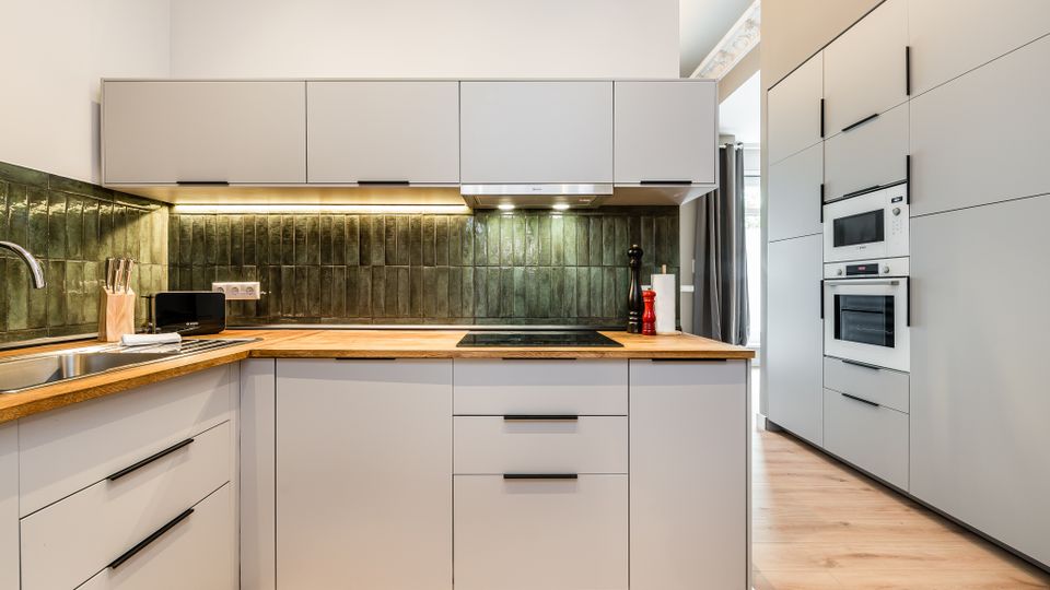 Moderne Einbauküche mit weißen, grifflosen Schränken, Holzarbeitsplatte und grün glasiertem Fliesenspiegel mit Unterbauleuchten. Rechts befindet sich ein hoher Schrankblock mit integriertem Backofen und Mikrowelle, der Raum hat hellen Holzboden und eine schlichte, helle Wandgestaltung.
