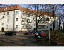 Wohngebäude mit weißer Fassade und rotem Ziegeldach an einer gepflasterten Straße, umgeben von Bäumen ohne Laub. Mehrere Balkone und Fenster prägen die Architektur, im Vordergrund sind parkende Autos, ein Roller und ein Straßenschild mit der Aufschrift „Epensteinstraße“ zu sehen.