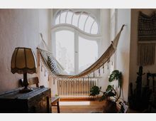 Heller Raum mit einem Bogenfenster, an dem eine Hängematte befestigt ist. Im Vordergrund stehen eine Tischlampe auf einer alten Holztruhe, ein Stuhl und mehrere Zimmerpflanzen auf einem Holzboden.