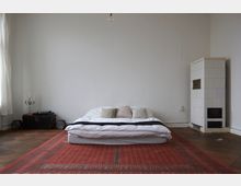 Minimalistisch eingerichtetes Schlafzimmer mit einem niedrigen Bett auf einem roten Teppich, einer weißen Wand, einem Kachelofen rechts und einem kleinen Beistelltisch mit einem Kerzenständer links. Ein hölzerner Boden ergänzt den Raum, beleuchtet von natürlichem Licht durch ein Fenster auf der linken Seite.