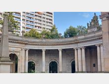 Steinernes halbrundes Denkmal mit Säulen und Reliefs, gekrönt von Skulpturen, umgeben von Bäumen; im Hintergrund ein modernes, hohes Wohngebäude mit Balkonen.