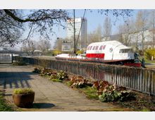 Ein befestigter Weg mit Pflanzenkübeln und Bodendeckern führt entlang eines schmiedeeisernen Geländers am Wasser. Im Hintergrund liegt ein fest vertäutes weiß-rotes Boot mit Plane, neben einem Schild, auf dem "Die Schiffskneipe" steht, sowie ein mehrstöckiges Bürogebäude und kahle Bäume.