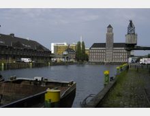 Industriehafen mit einem breiten Wasserkanal, umgeben von historischen Backsteingebäuden und moderneren Bürogebäuden im Hintergrund. Auf der rechten Seite stehen ein alter Kran und gelb-grüne Poller entlang einer gepflasterten Uferpromenade.