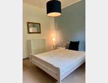 Schlafzimmer mit hellem Holzbett und weißer Bettwäsche, blaugrauer Akzentwand und Teppichboden. An der Wand hängen ein gerahmter Spiegel über einem weißen Schrank; neben dem Bett stehen ein kleiner Nachttisch und eine hohe Stehlampe, ergänzt durch eine schwarze Deckenleuchte.