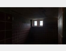 Ein dunkler Raum mit rotbraunen Fliesen an den Wänden und einem kleinen, vergitterten Fenster, das schwaches Licht hereinlässt. Die Decke wirkt niedrig und teilweise beschädigt.