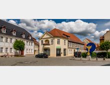 Kleiner Platz mit gepflasterter Straße, umgeben von historischen Gebäuden mit roten Ziegeldächern und Fachwerkelementen. Eine blaue Skulptur mit einem gelben Engel steht zentral, umgeben von einigen geparkten Autos und Fahrrädern.