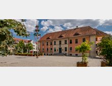 Historisches Gebäude mit orangefarbener Fassade und Ziegeldach, zentral auf einem gepflasterten Platz gelegen. Im Vordergrund ein geschmückter Maibaum, umgeben von Blumenkübeln und Grünpflanzen; im Hintergrund weitere Gebäude und blauer Himmel mit wenigen Wolken.